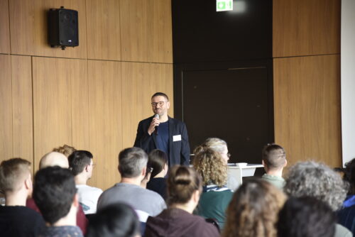 Person mit Mikrofon vor Publikum in einem Raum mit Holzvertäfelung und Lautsprecher an der Wand