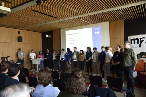 Gruppe von Personen steht vor Publikum in einem Raum mit Holzpaneelen und einer Projektionsfläche mit Text zur Vorstellung von Impulsen