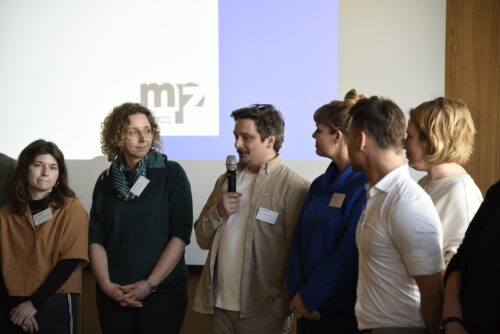 Sechs Personen stehen nebeneinander vor einer Wand mit projiziertem mpz-Logo, eine Person spricht in ein Mikrofon