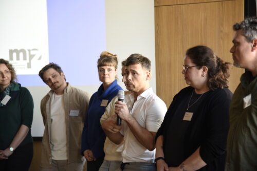 Sechs Personen stehen nebeneinander vor einer Wand mit projiziertem mpz-Logo, eine Person spricht in ein Mikrofon