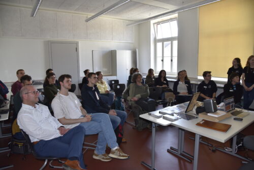 Workshops - Gruppe von Personen sitzt in einem hellen Seminarraum mit großen Fenstern und blickt auf einen Tisch mit Laptops und Präsentationsmaterial