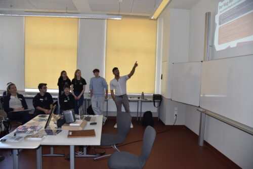 Gruppe von Personen in einem Seminarraum mit einem Vortragenden, der auf eine projizierte Präsentation an der Wand zeigt